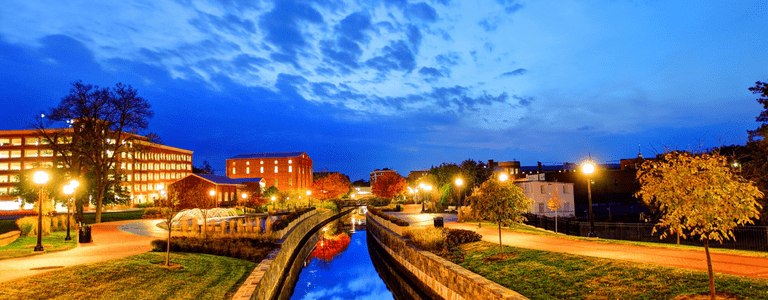 A scenic evening view of a canal lined with trees, illuminated streetlights, and historic brick buildings under a vibrant blue sky