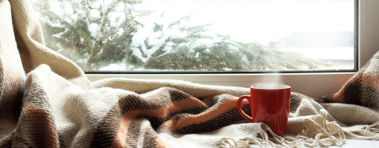 A cozy scene with a warm blanket and a steaming red mug by a window, overlooking snow-covered trees outside